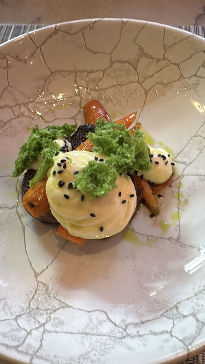 Plated gourmet dish with roasted carrots, creamy sauce, green garnish, and black sesame seeds on a decorative white plate.