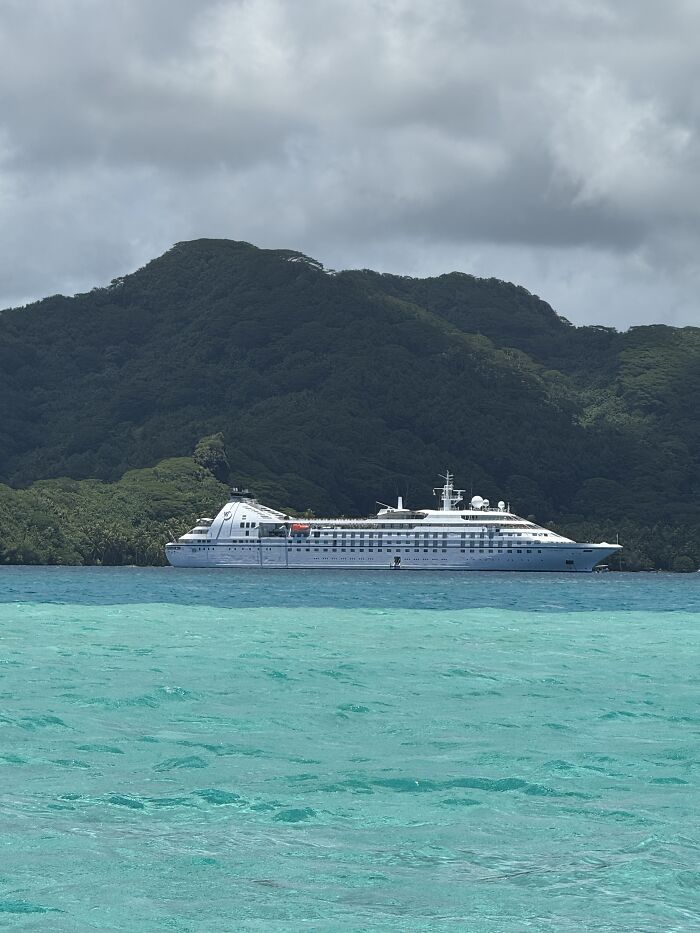 Large white cruise ship sails through vibrant shades of blue water near lush green mountains in French Polynesia under cloudy sky.