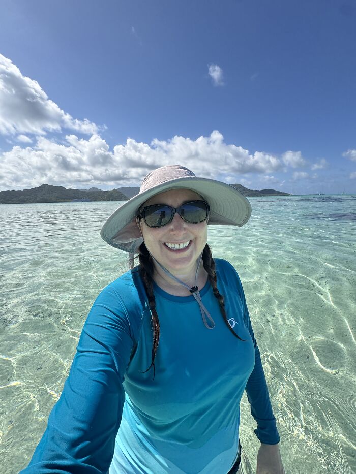 Woman in blue shirt and hat smiling in clear shallow water under blue sky in French Polynesia ocean.