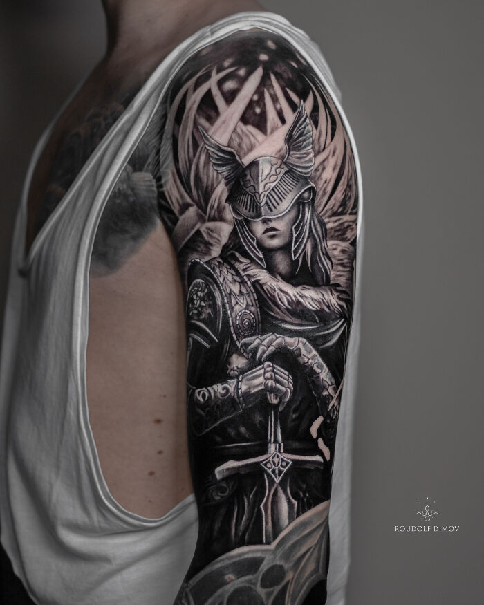 Black and grey realistic tattoo of a warrior with a helmet and wings, holding a sword, on a person's upper arm.