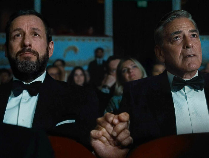Two men in tuxedos holding hands in a theater audience during the Academy releases 2026 Oscar nominations event.