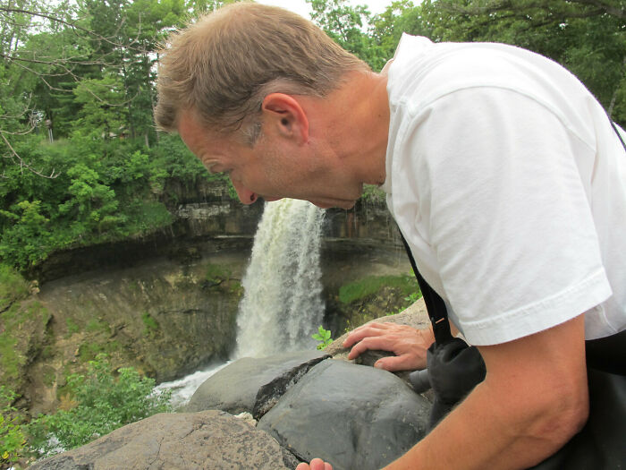 Man in white shirt leaning over rocks with waterfall appearing like funny confusing picture of water pouring from his mouth outdoors