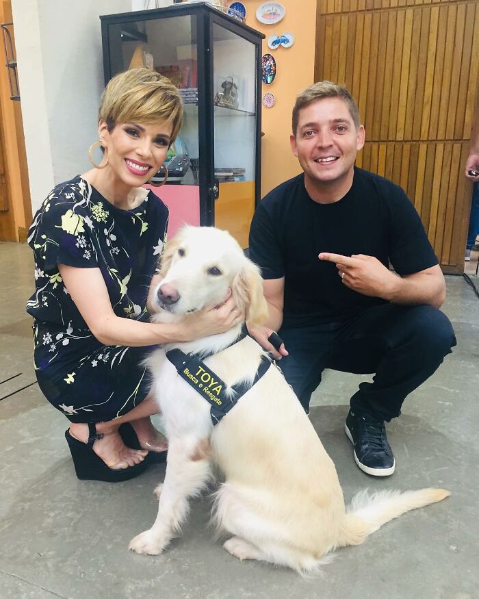 Two people smiling while posing with a golden retriever dog wearing a harness indoors, symbolizing human-animal bonding. Two people smiling while posing with a golden retriever dog wearing a harness indoors, symbolizing human-animal bonding.