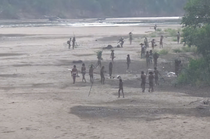 Uncontacted Amazonian tribe members on riverbank holding spears amid dense forest in rare never-before-seen footage.