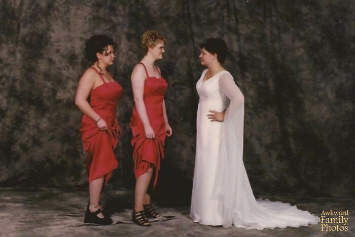Bride in a white dress facing two bridesmaids in red dresses, awkward wedding photo with unusual poses against a dark backdrop.