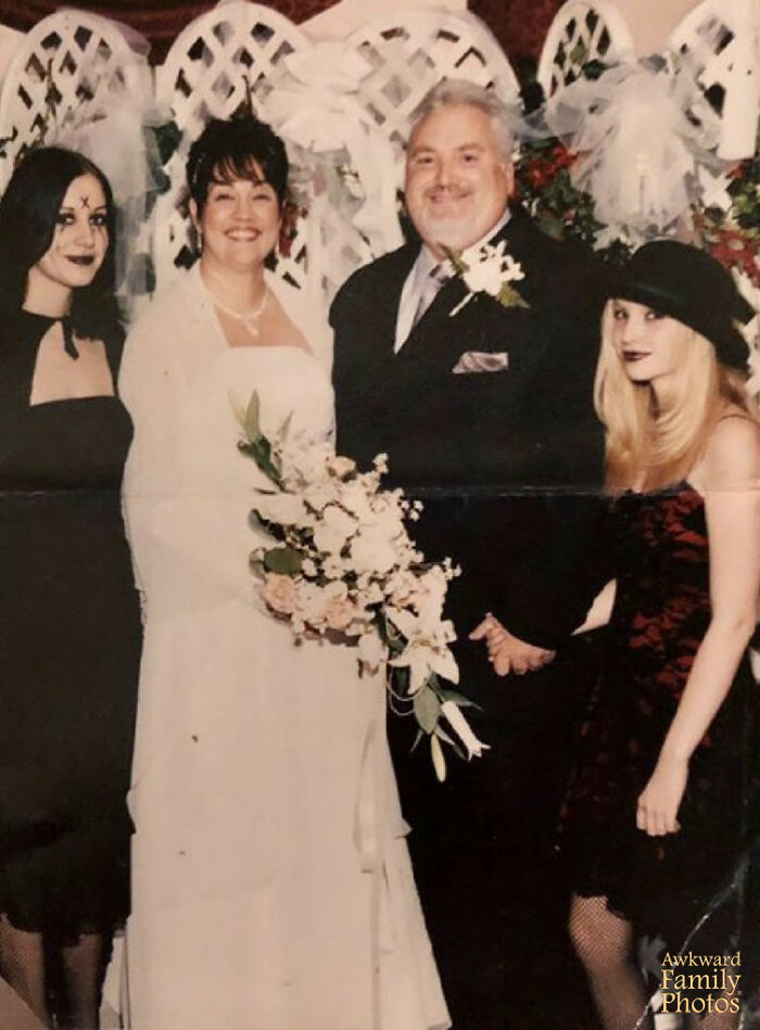 Bride and groom smiling with two guests in gothic outfits at an awkward wedding photo with contrasting styles.