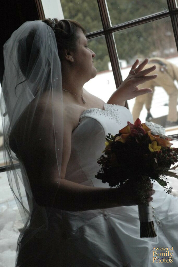 Bride in wedding dress holding bouquet by window while person outside creates an awkward wedding photo moment.