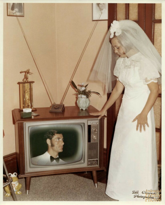 Bride in a vintage wedding dress looking at a groom’s face on an old TV in an awkward wedding photo moment.