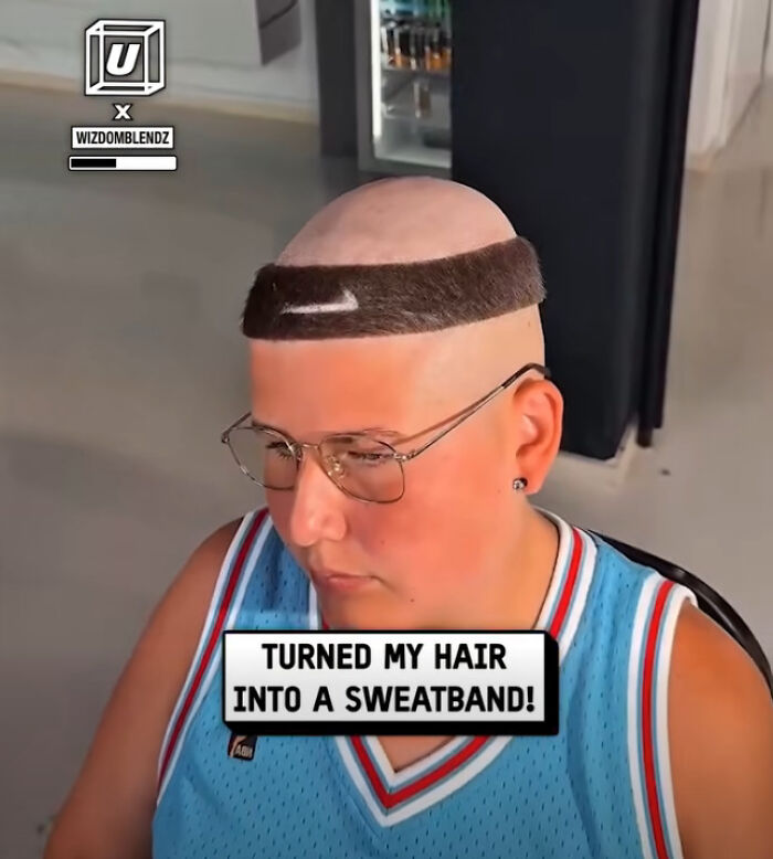 Man with disastrous hairstyle shaved bald except for a thick hair strip designed like a sweatband, wearing glasses and a blue jersey.