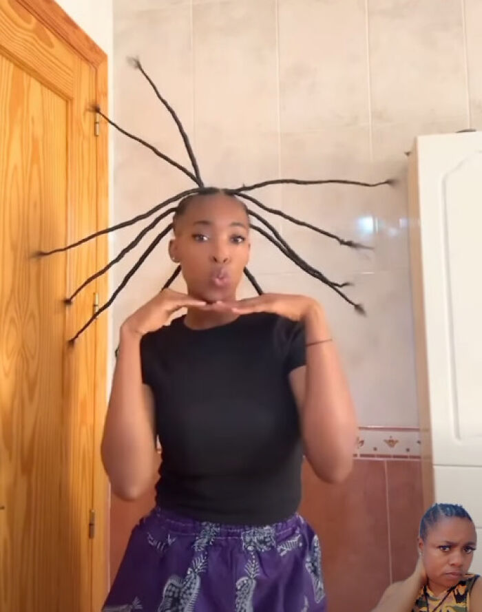 Woman with disastrous hairstyles styled into long braids sticking out from her head, posing indoors in a casual outfit.