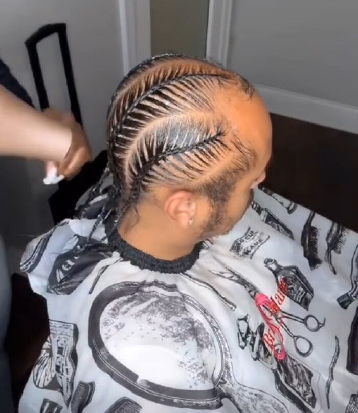 Man with a unique and intricate braided hairstyle sitting in a barber chair wearing a printed haircut cape.
