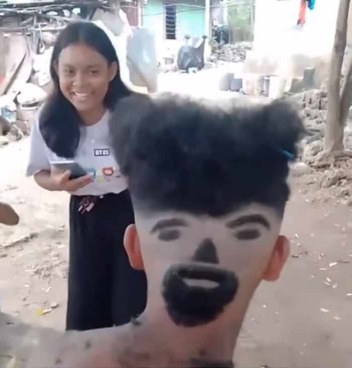 Young man with a disastrous hairstyle featuring a face design shaved into the back of his head, while a girl smiles nearby outdoors.