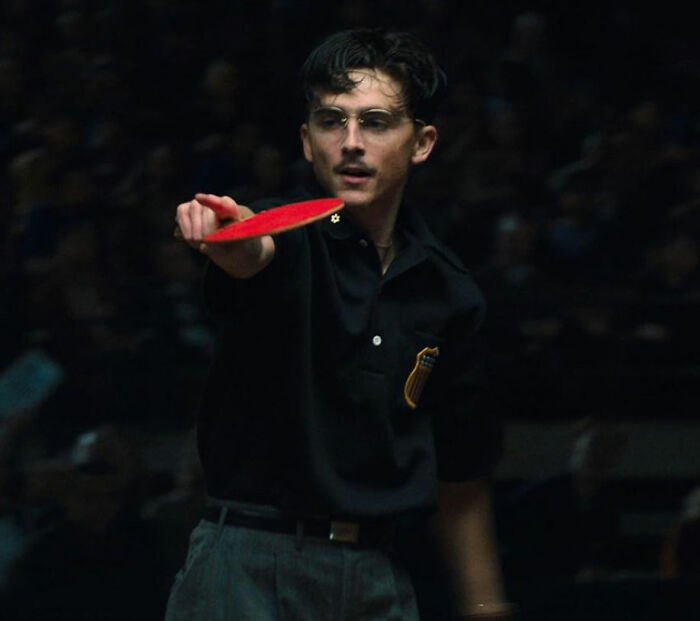Young man playing table tennis intensely, showcasing focus and skill in a dimly lit indoor setting for Oscar nominations.