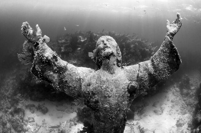 Underwater photo of a weathered statue with arms raised surrounded by marine life and coral reef environment.