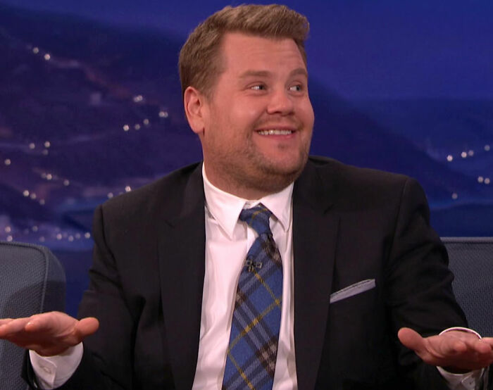 Man in a suit and patterned tie smiling and gesturing during a talk show about movie set workers and diva stars.