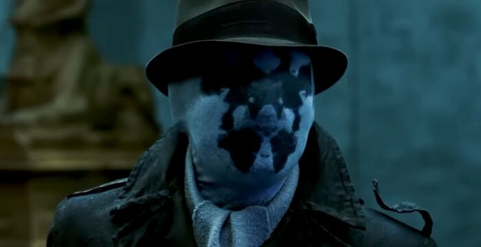 Rorschach from Watchmen wearing a fedora and trench coat, representing iconic famous villains in dark lighting.