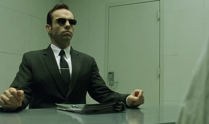 Man in black suit and sunglasses sitting at table with folder, evoking themes of famous villains and pure evil discussions.