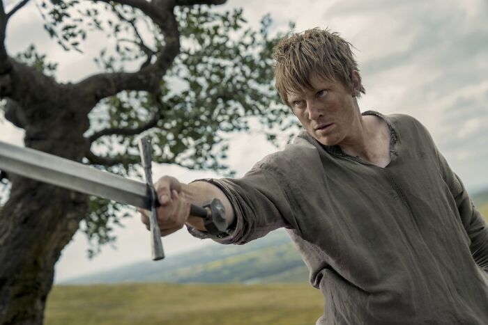 Hedge knight in Knight of the Seven Kingdoms holding sword outdoors with focused expression and rustic background.