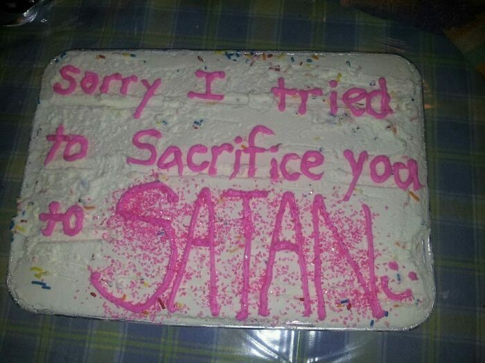 Funny cake with misspelled pink icing text expressing apology for trying to sacrifice someone to Satan, humorous emotion.