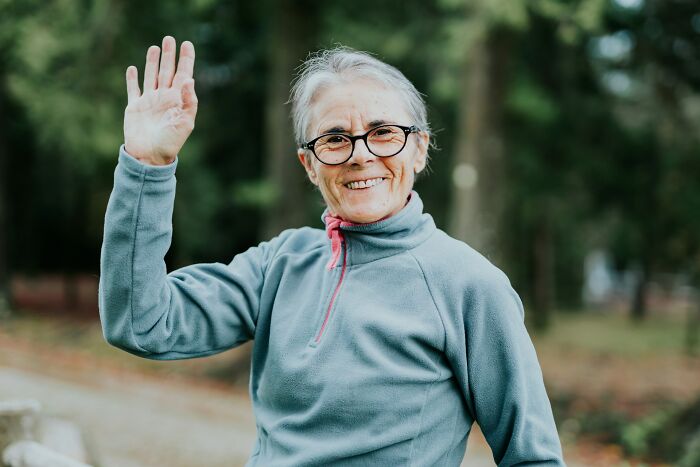 Elderly woman in glasses wearing a gray pullover, smiling and waving hand, illustrating things normal in America but offensive elsewhere.