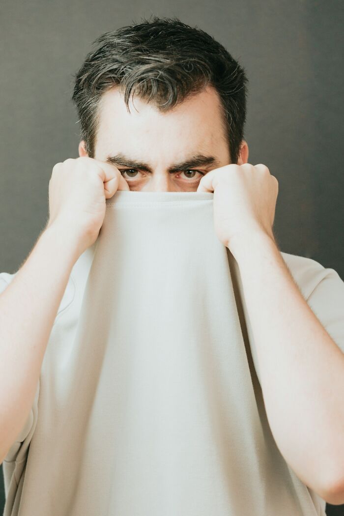Man hiding face behind shirt, showing expression of panic and anxiety related to accidental texts situation.
