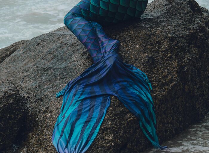 Colorful mermaid tail resting on a rocky shore near water, illustrating a fantasy scene away from worst coworkers at work.