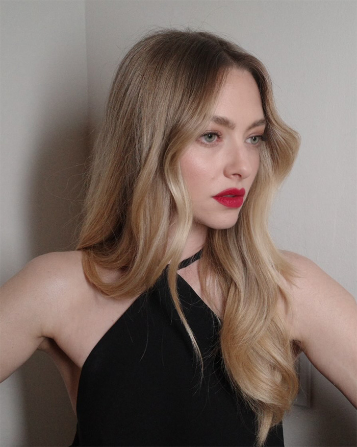 Amanda Seyfried with long blonde hair and red lipstick, wearing a black top, showing a serious expression.