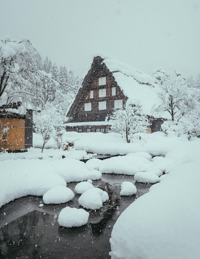 Snow-covered traditional house and trees by a partially frozen stream, capturing cozy winter scenery to enjoy indoors.
