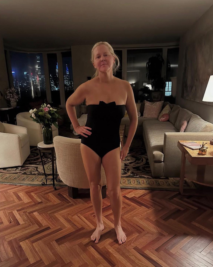 Amy Schumer in a black swimsuit standing in a living room with city lights visible through the windows at night. Amy Schumer in a black swimsuit standing in a living room with city lights visible through the windows at night.