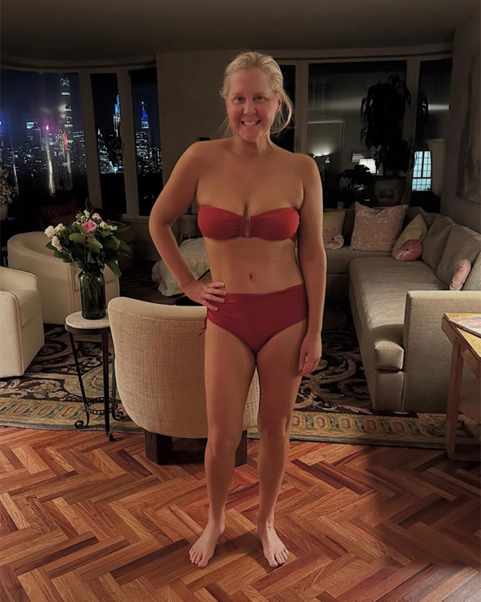 Amy Schumer smiling in a red bikini indoors with city lights visible through the window at night. Amy Schumer smiling in a red bikini indoors with city lights visible through the window at night.