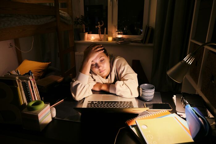 Tired young woman working late at desk with laptop and notebooks, reflecting on adult goals in a dimly lit room.