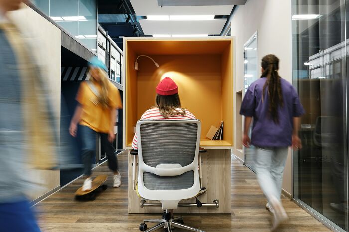 Person in a red beanie working alone in a small office pod while coworkers move past in a busy modern workplace.