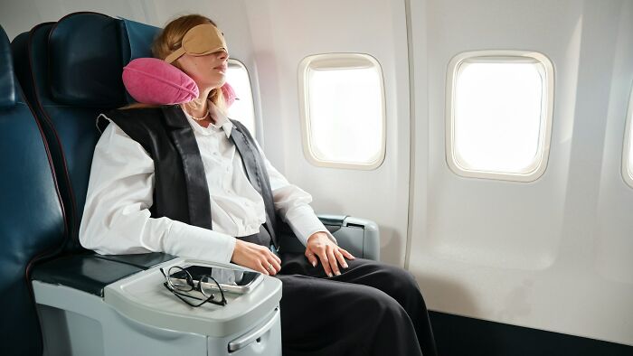 Woman with neck pillow and eye mask seated in airplane window seat, appearing relaxed during the flight experience.