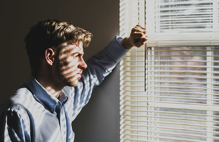 Young man in blue shirt looking thoughtfully through window blinds, contemplating landlord profits and relationship demands. Young man in blue shirt looking thoughtfully through window blinds, contemplating landlord profits and relationship demands.