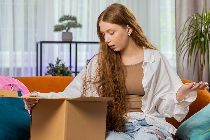 Young woman with long hair sitting on a couch, looking confused while packing a cardboard box in a living room setting Young woman with long hair sitting on a couch, looking confused while packing a cardboard box in a living room setting