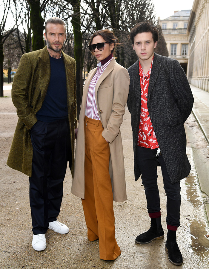 Victoria Beckham standing outdoors with two men, wearing sunglasses and a beige coat in a tense exchange amid feud. Victoria Beckham standing outdoors with two men, wearing sunglasses and a beige coat in a tense exchange amid feud.