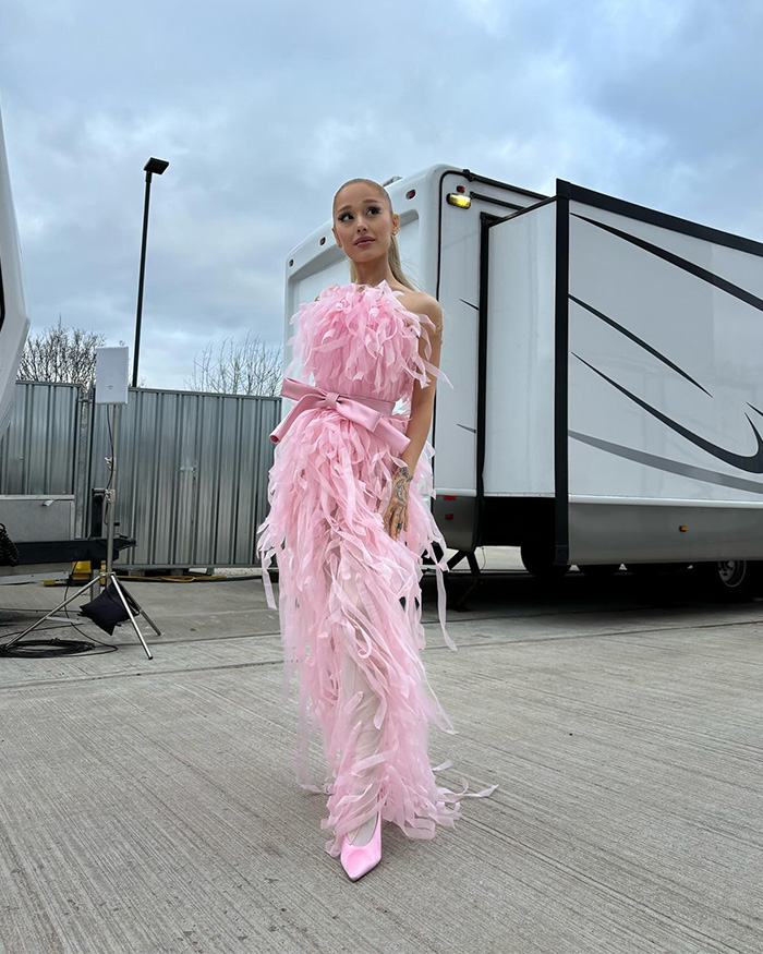 Ariana Grande wearing a pink feathered gown and high heels standing outdoors near trailers showcasing her transformation.