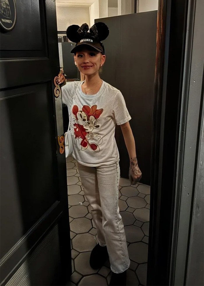 Young woman wearing a Mickey Mouse T-shirt and cap stands in a doorway, showcasing Ariana Grande's transformation in new pics. Young woman wearing a Mickey Mouse T-shirt and cap stands in a doorway, showcasing Ariana Grande's transformation in new pics.