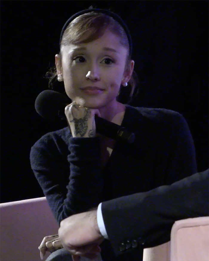 Ariana Grande holding a microphone, wearing a black top and headband, showing her tattooed hand during a discussion. Ariana Grande holding a microphone, wearing a black top and headband, showing her tattooed hand during a discussion.