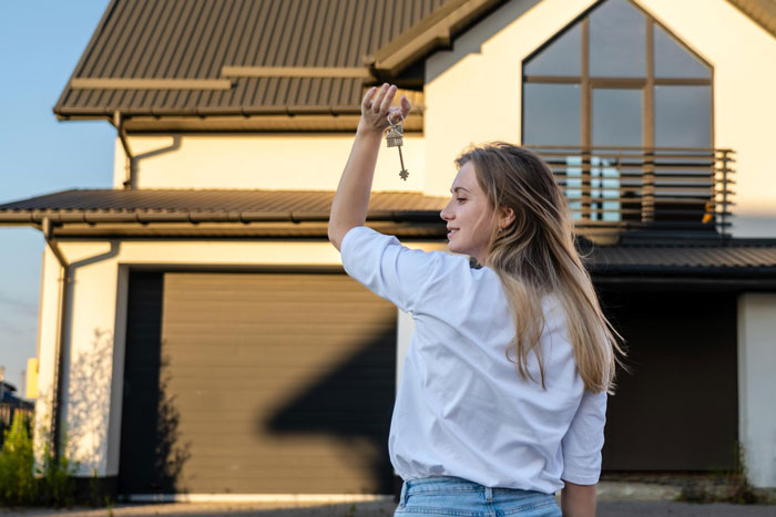 Young woman holding house keys in front of suburban home, parents house payment concept Young woman holding house keys in front of suburban home, parents house payment concept