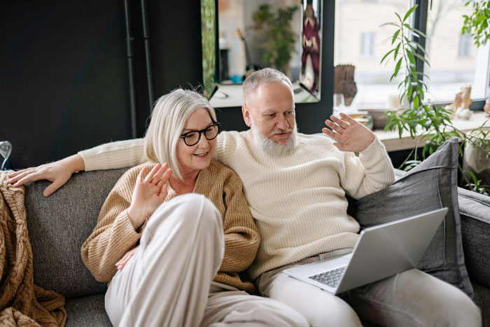 Senior parents sitting on couch waving at laptop during video call about parents house payment