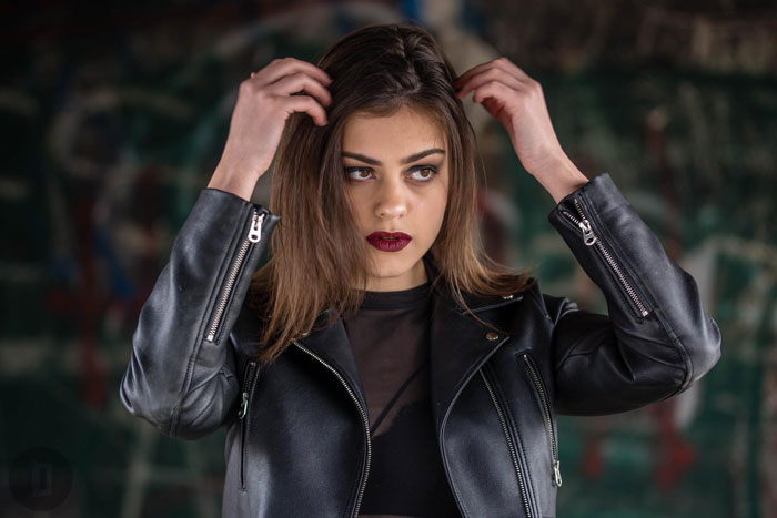 Young woman in a black leather jacket adjusting hair, posing confidently with a camera-ready look in an urban family setting.