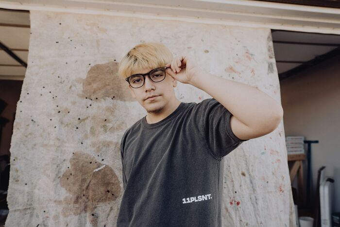 Young man with blonde hair and glasses adjusting frames, representing cultural differences in America and offensive gestures elsewhere.