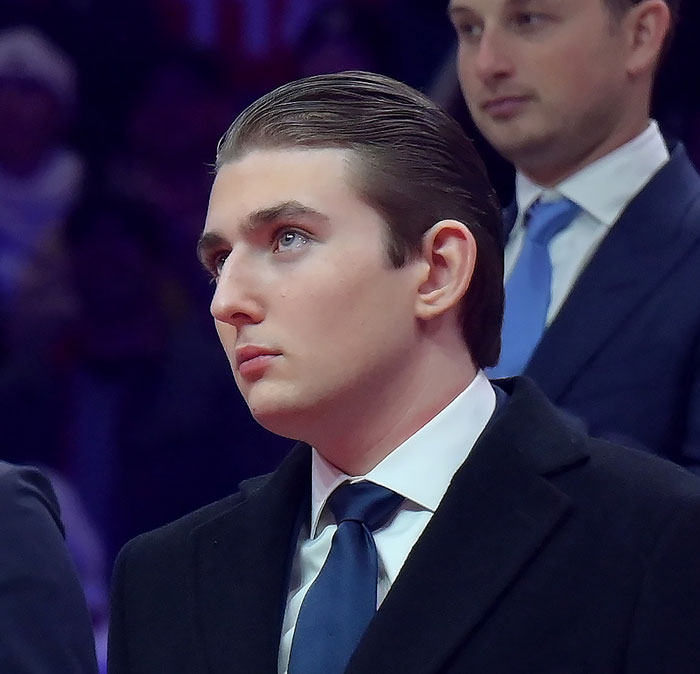 Barron Trump vestindo terno e gravata, olhando atentamente durante um evento formal ao lado de outro homem.