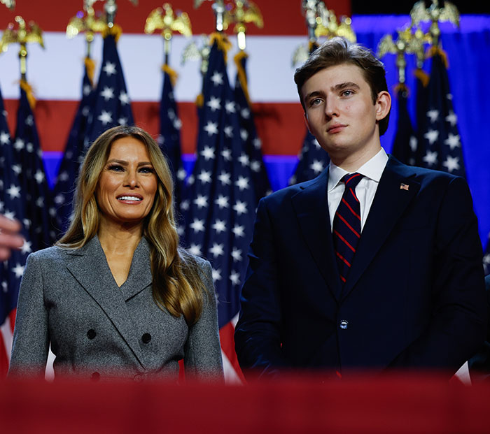 Barron Trump ao lado de uma mulher com bandeiras americanas ao fundo durante um evento formal. Barron Trump ao lado de uma mulher com bandeiras americanas ao fundo durante um evento formal.