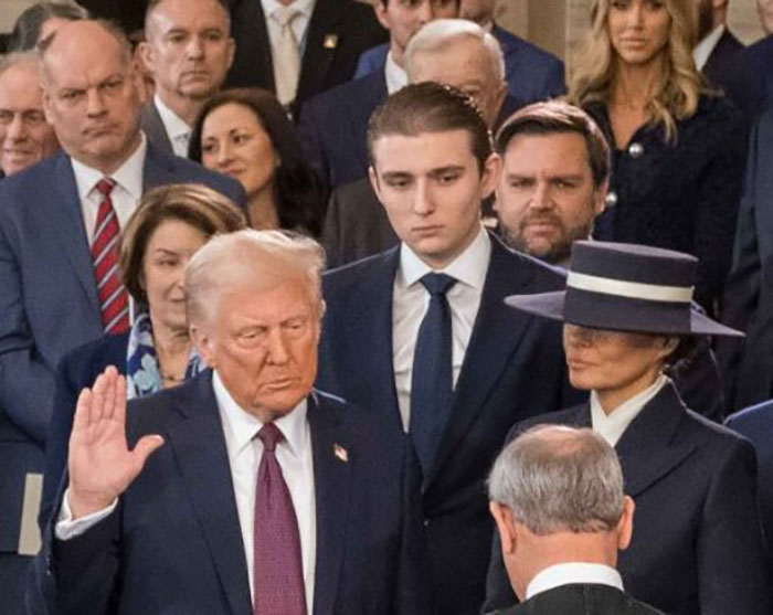 Barron Trump atrás de Donald Trump durante um evento formal, parecendo sério e atento no meio da multidão.