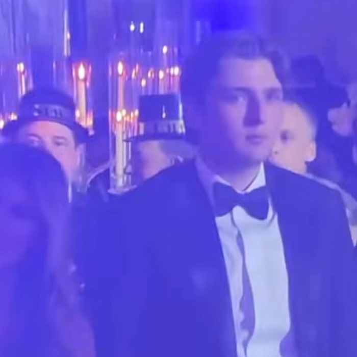 Barron Trump in a tuxedo at a New Year’s Eve party with a serious expression amid dim blue lighting. Barron Trump in a tuxedo at a New Year’s Eve party with a serious expression amid dim blue lighting.
