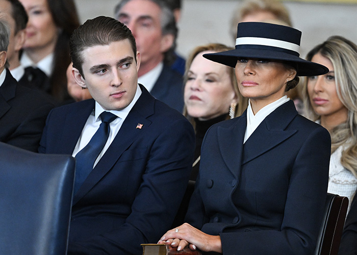Barron Trump and Melania Trump seated, dressed formally, with a serious expression amid a crowd at a public event. Barron Trump and Melania Trump seated, dressed formally, with a serious expression amid a crowd at a public event.