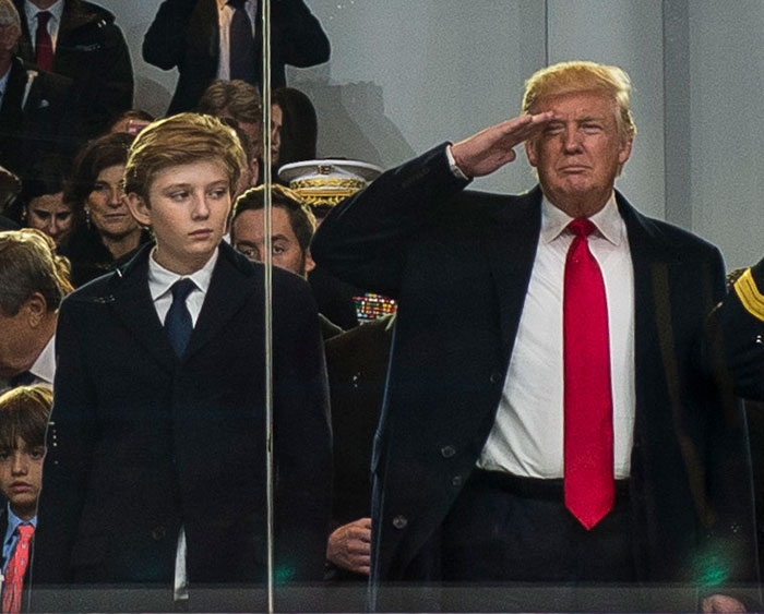 Barron Trump ao lado de um homem fazendo continência durante um evento formal com pessoas sentadas ao fundo. Barron Trump ao lado de um homem fazendo continência durante um evento formal com pessoas sentadas ao fundo.