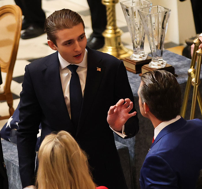 Barron Trump falando com duas pessoas de terno em um evento formal, conversando durante uma discussão por chamada de emergência.